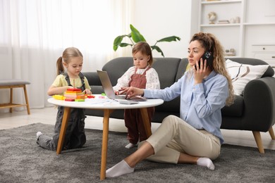 Photo of Single mother talking on smartphone and working with laptop while her daughters playing indoors