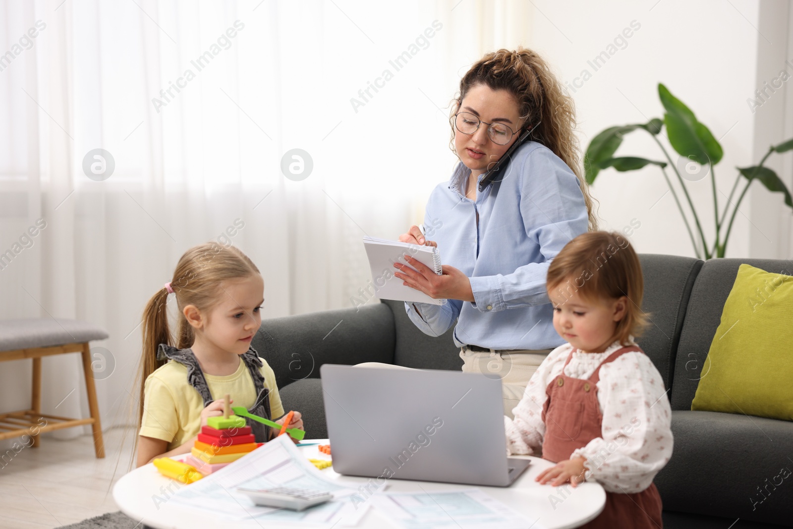 Photo of Work-family balance. Single mother taking notes while talking on smartphone and her daughters at home