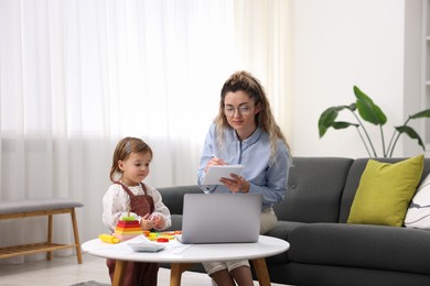 Photo of Work-family balance. Single mother taking notes while her daughter playing with toy indoors