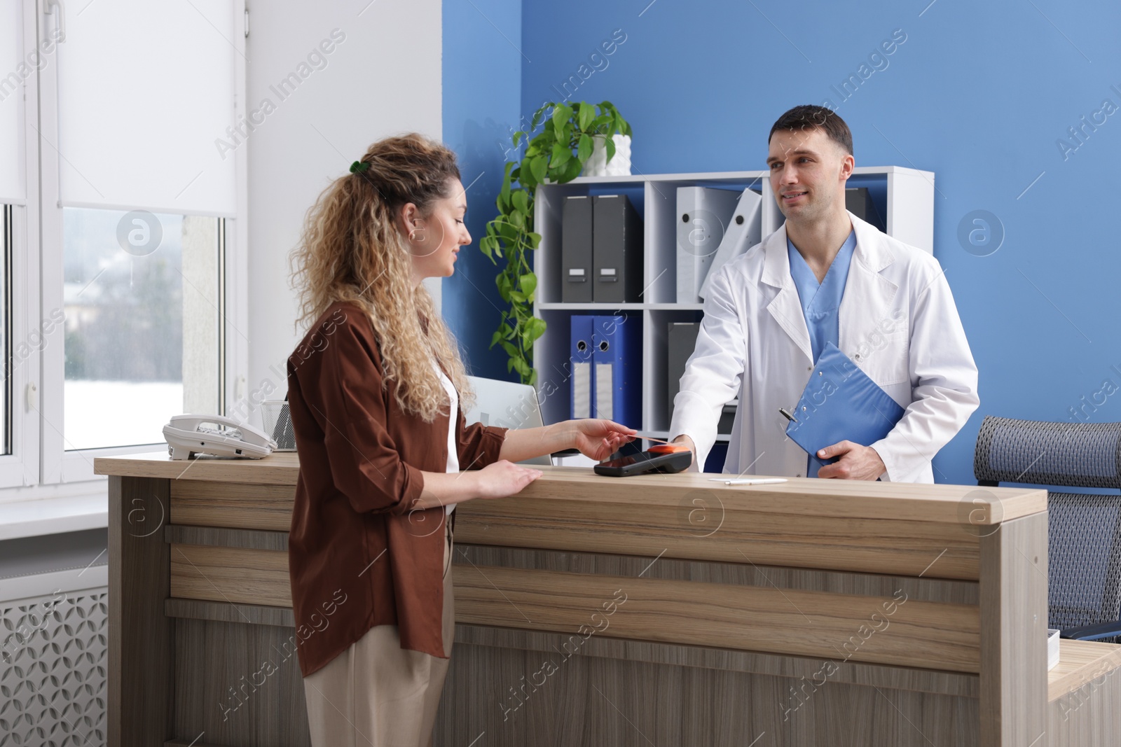 Photo of Smiling receptionist taking payment from patient via terminal at counter in clinic