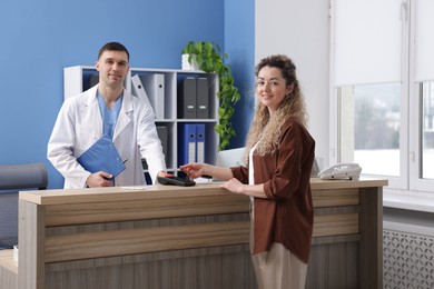 Photo of Smiling receptionist taking payment from patient via terminal at counter in clinic