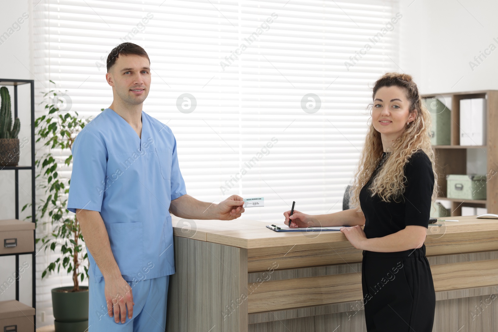 Photo of Smiling patient filling document and receptionist with medical insurance card at counter in clinic