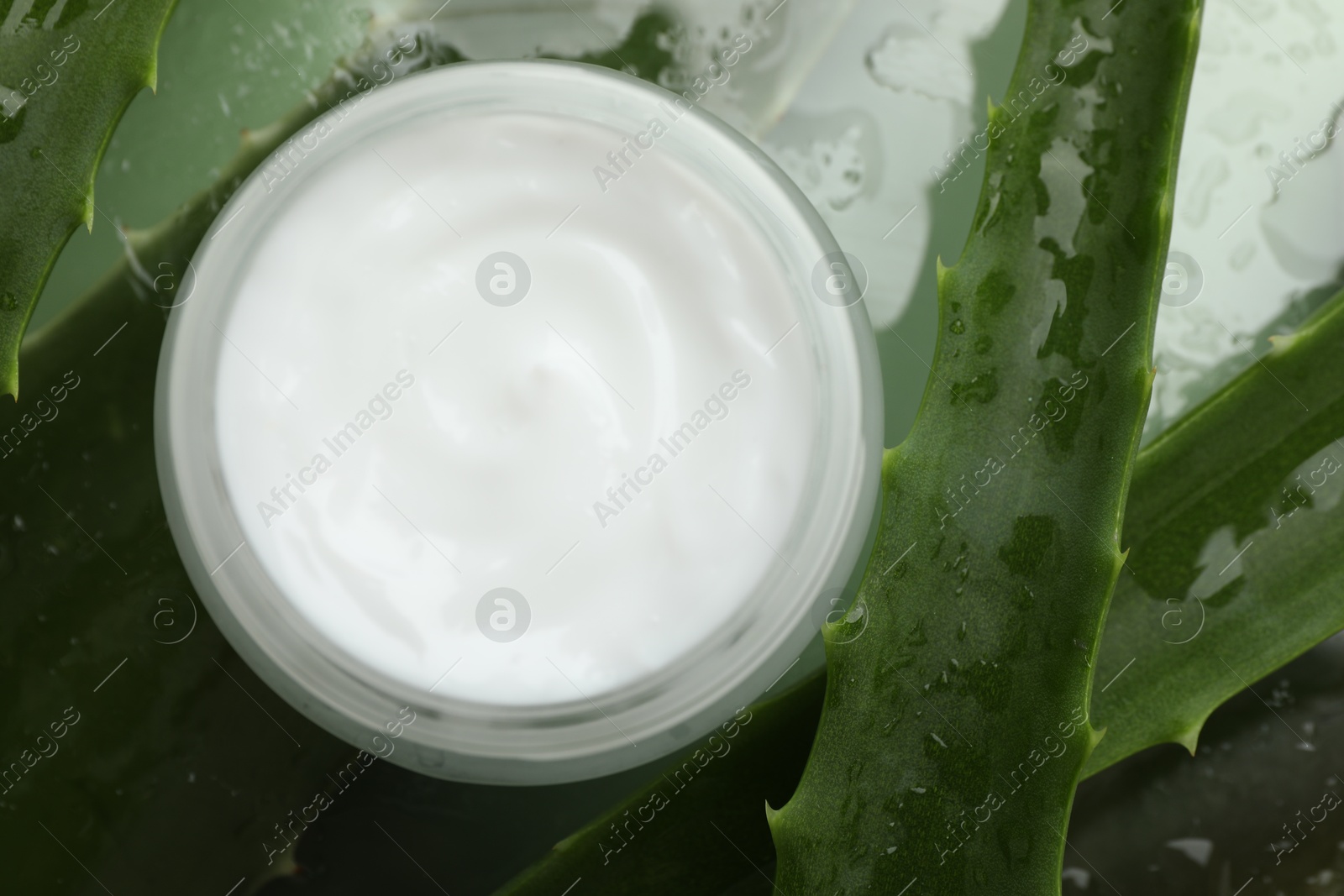 Photo of Moisturizing cream with extract of aloe vera and wet leaves on mirror surface, flat lay