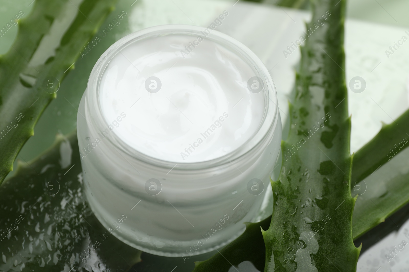 Photo of Moisturizing cream with extract of aloe vera and wet leaves on mirror surface, closeup