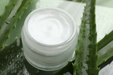 Moisturizing cream with extract of aloe vera and wet leaves on mirror surface, closeup