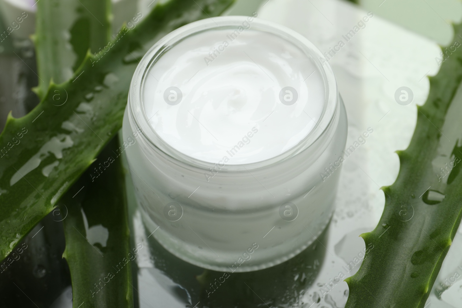 Photo of Moisturizing cream with extract of aloe vera and wet leaves on mirror surface, closeup