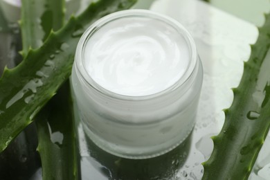 Moisturizing cream with extract of aloe vera and wet leaves on mirror surface, closeup