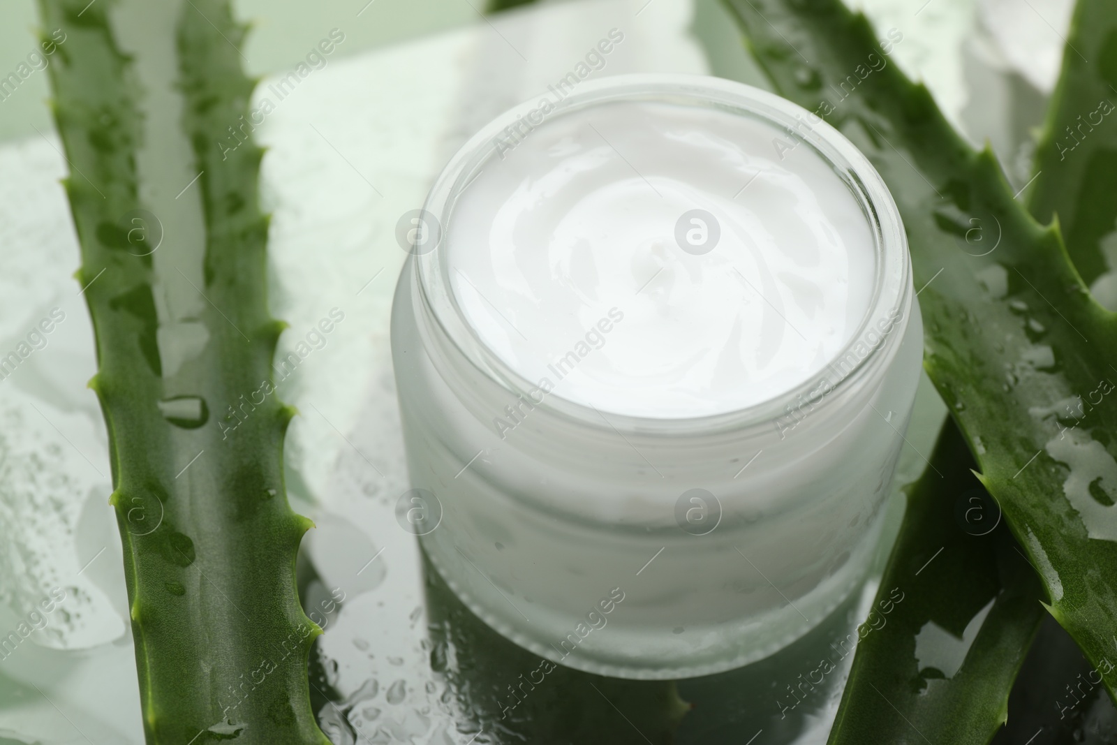 Photo of Moisturizing cream with extract of aloe vera and wet leaves on mirror surface, closeup