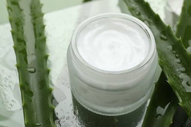 Moisturizing cream with extract of aloe vera and wet leaves on mirror surface, closeup