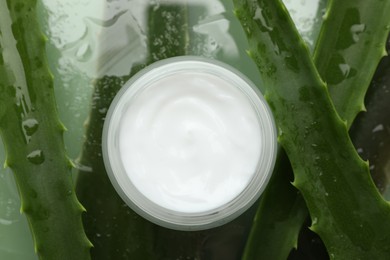 Photo of Moisturizing cream with extract of aloe vera and wet leaves on mirror surface, flat lay