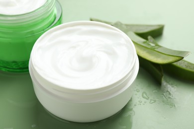 Moisturizing cream with extract of aloe vera, cut leaves and water drops on green background, closeup