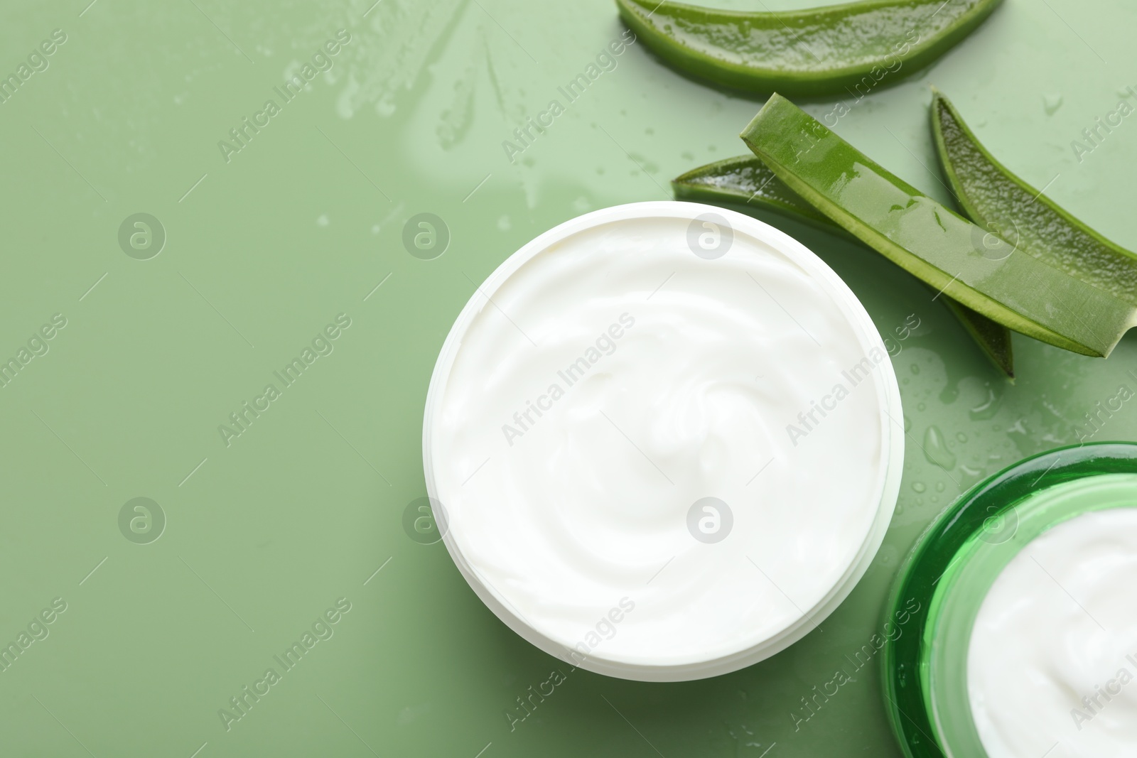 Photo of Moisturizing cream with extract of aloe vera, cut leaves and water drops on green background, flat lay. Space for text