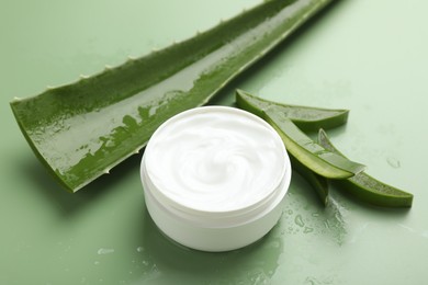 Photo of Moisturizing cream with extract of aloe vera, cut leaves and water drops on green background, closeup