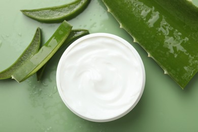 Moisturizing cream with extract of aloe vera, cut leaves and water drops on green background, flat lay