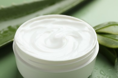 Photo of Moisturizing cream with extract of aloe vera, cut leaves and water drops on green background, closeup