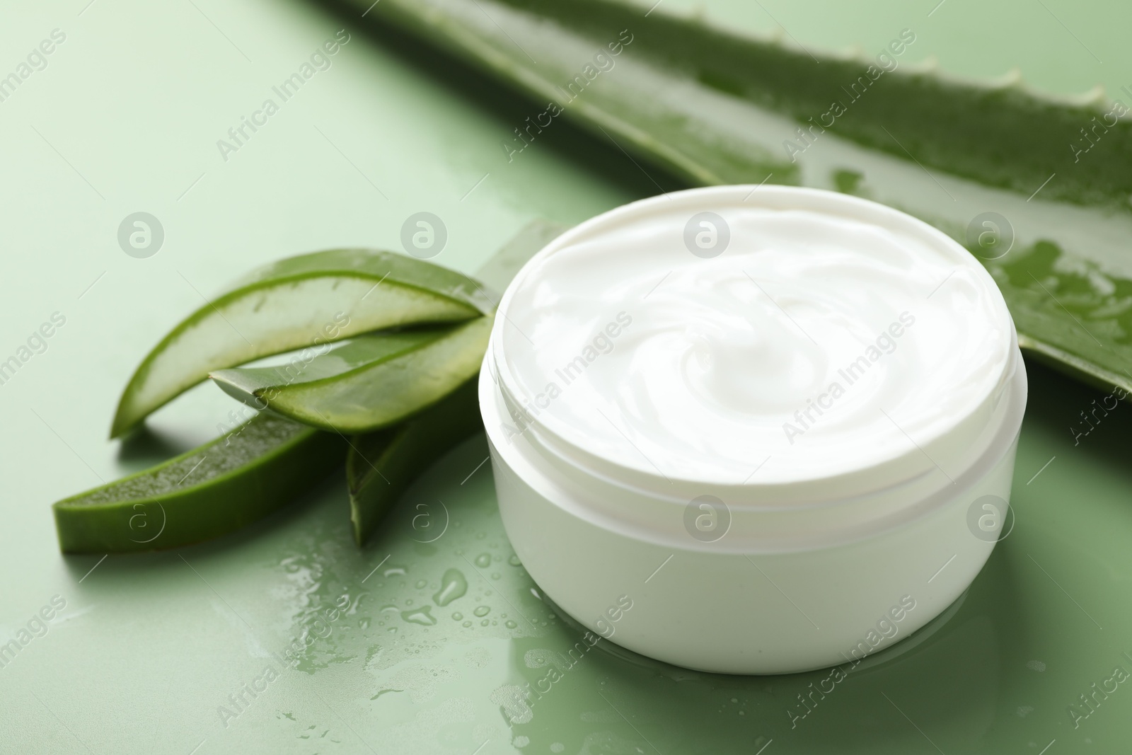 Photo of Moisturizing cream with extract of aloe vera, cut leaves and water drops on green background, closeup