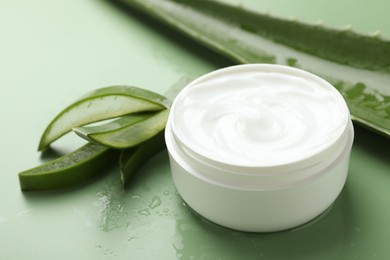 Photo of Moisturizing cream with extract of aloe vera, cut leaves and water drops on green background, closeup