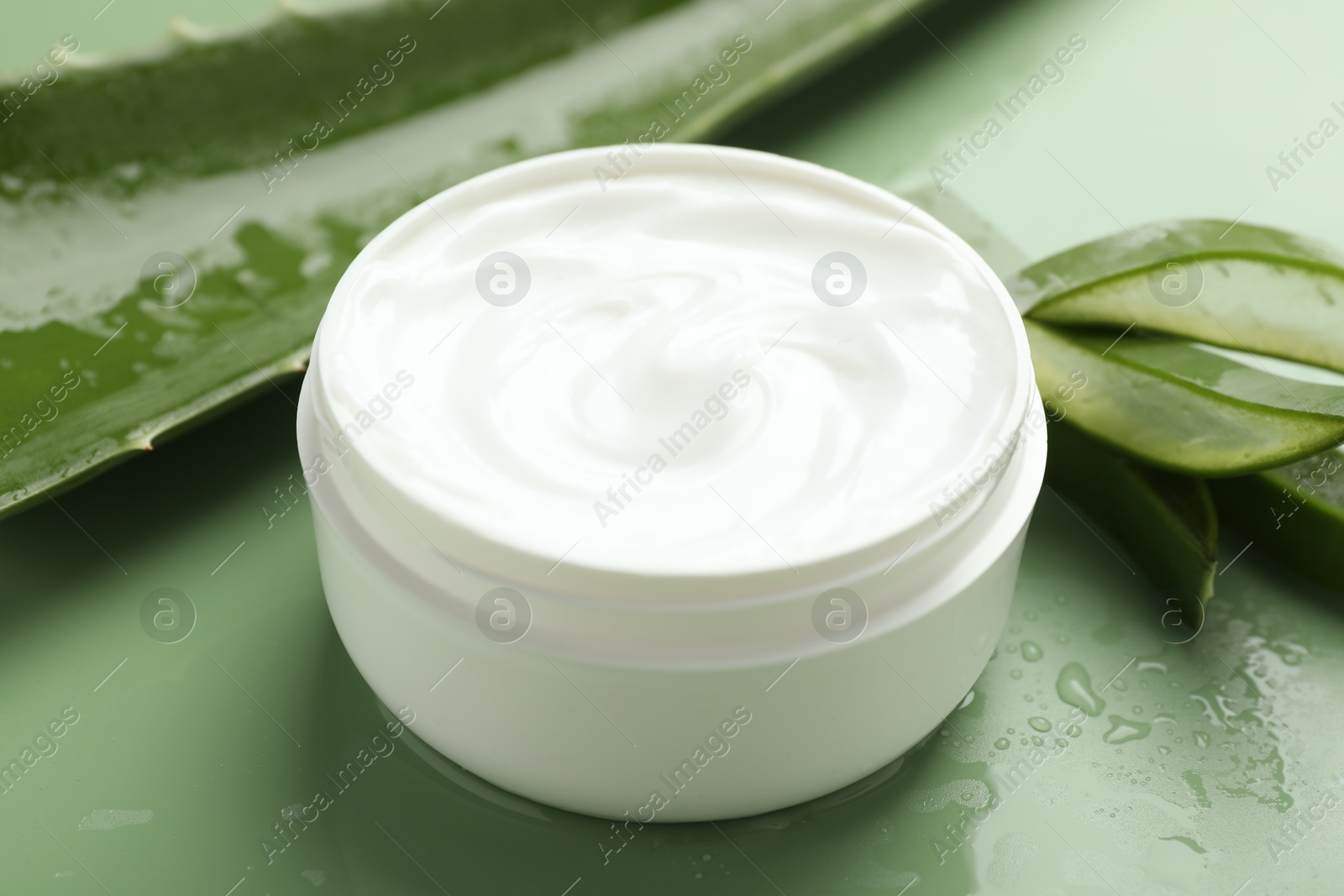 Photo of Moisturizing cream with extract of aloe vera, cut leaves and water drops on green background, closeup