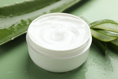 Moisturizing cream with extract of aloe vera, cut leaves and water drops on green background, closeup