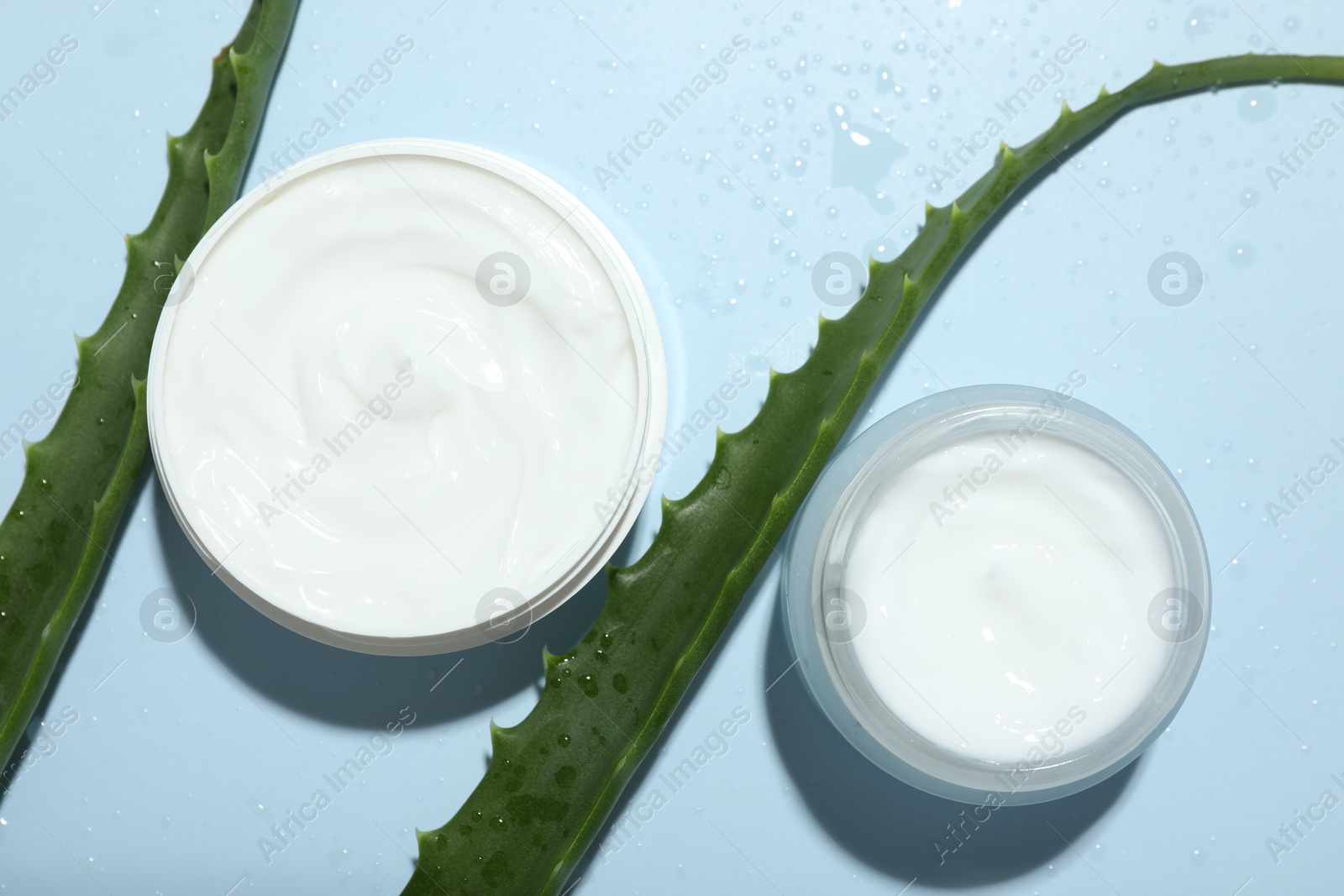 Photo of Moisturizing cream with extract of aloe vera, cut leaves and water drops on light blue background, flat lay