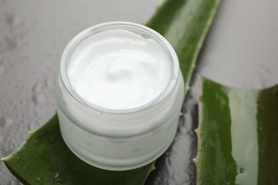 Photo of Moisturizing cream with extract of aloe vera in jar, cut leaves and water drops on grey background, closeup