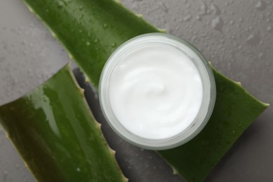 Photo of Moisturizing cream with extract of aloe vera in jar, cut leaves and water drops on grey background, flat lay