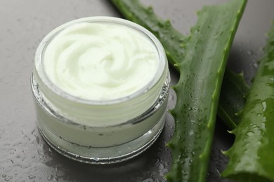 Photo of Moisturizing cream with extract of aloe vera in jar, cut leaves and water drops on grey background, closeup