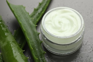 Photo of Moisturizing cream with extract of aloe vera in jar, cut leaves and water drops on grey background, closeup