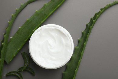 Photo of Moisturizing cream with extract of aloe vera in jar and cut leaves on grey background, flat lay