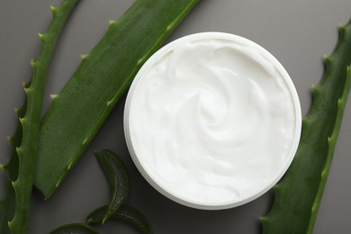 Photo of Moisturizing cream with extract of aloe vera in jar and cut leaves on grey background, flat lay