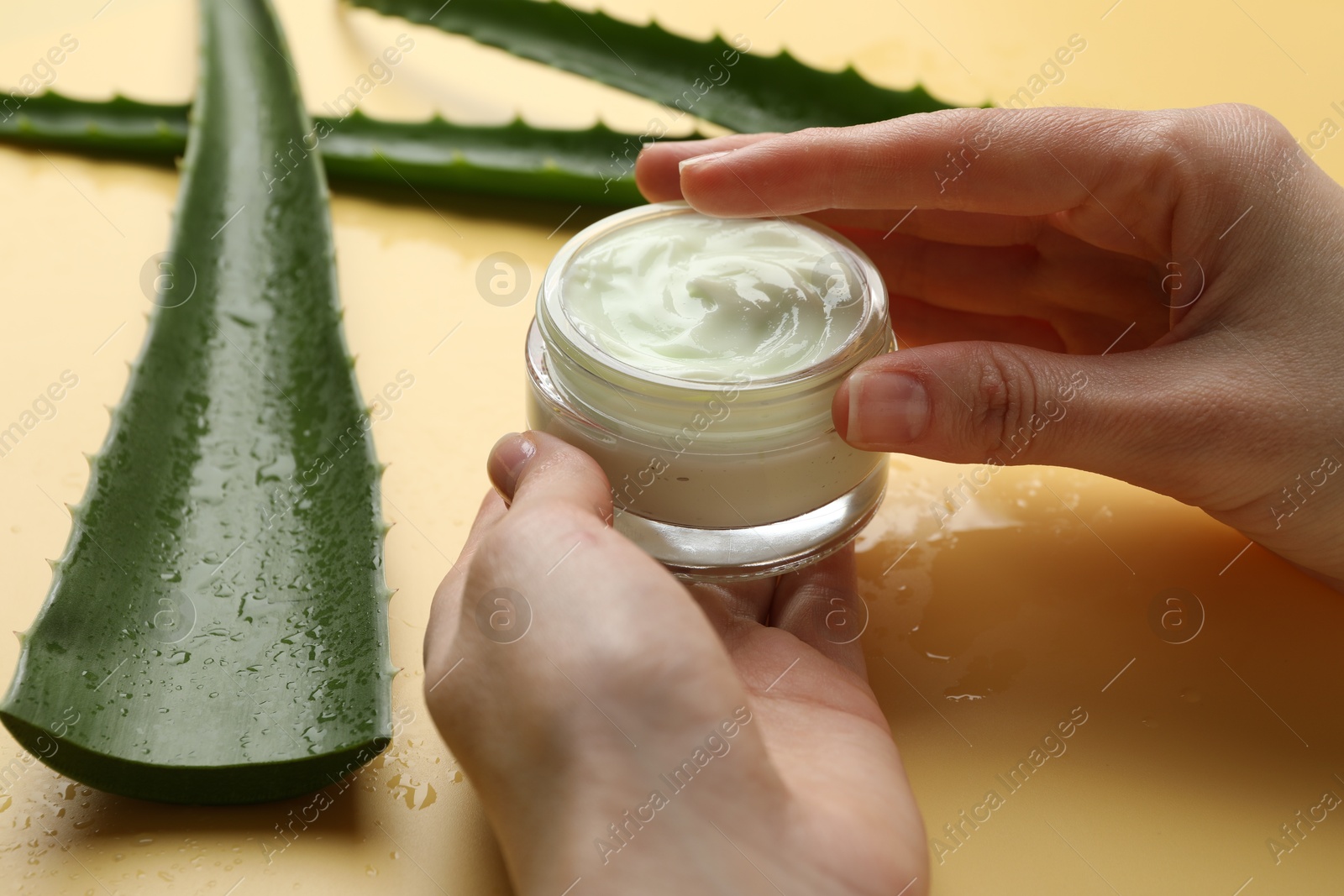 Photo of Woman with moisturizing cream and wet aloe vera leaves on yellow background, closeup