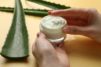 Photo of Woman with moisturizing cream and wet aloe vera leaves on yellow background, closeup