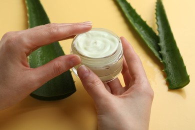 Woman with moisturizing cream and wet aloe vera leaves on yellow background, closeup