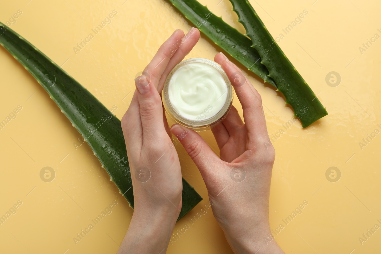 Photo of Woman with moisturizing cream and wet aloe vera leaves on yellow background, top view