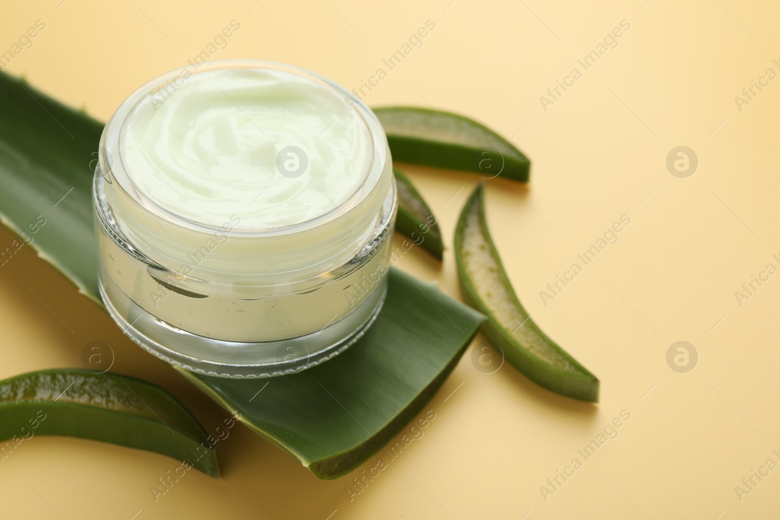 Photo of Moisturizing cream with extract of aloe vera in jar and cut leaves on yellow background, closeup