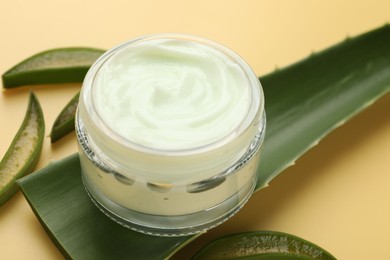 Photo of Moisturizing cream with extract of aloe vera in jar and cut leaves on yellow background, closeup