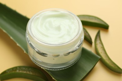 Photo of Moisturizing cream with extract of aloe vera in jar and cut leaves on yellow background, closeup