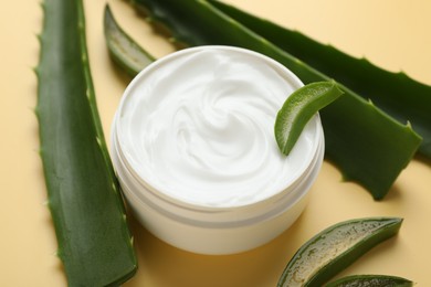 Photo of Moisturizing cream with extract of aloe vera in jar and cut leaves on yellow background, closeup