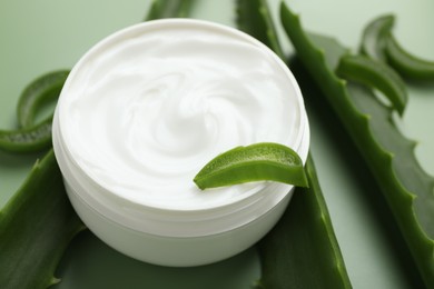 Moisturizing cream with extract of aloe vera in jar and cut leaves on green background, closeup