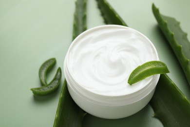 Moisturizing cream with extract of aloe vera in jar and cut leaves on green background, closeup