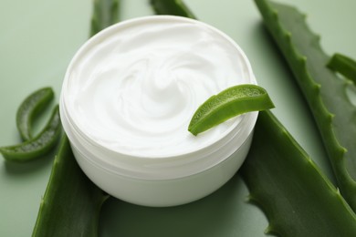 Photo of Moisturizing cream with extract of aloe vera in jar and cut leaves on green background, closeup