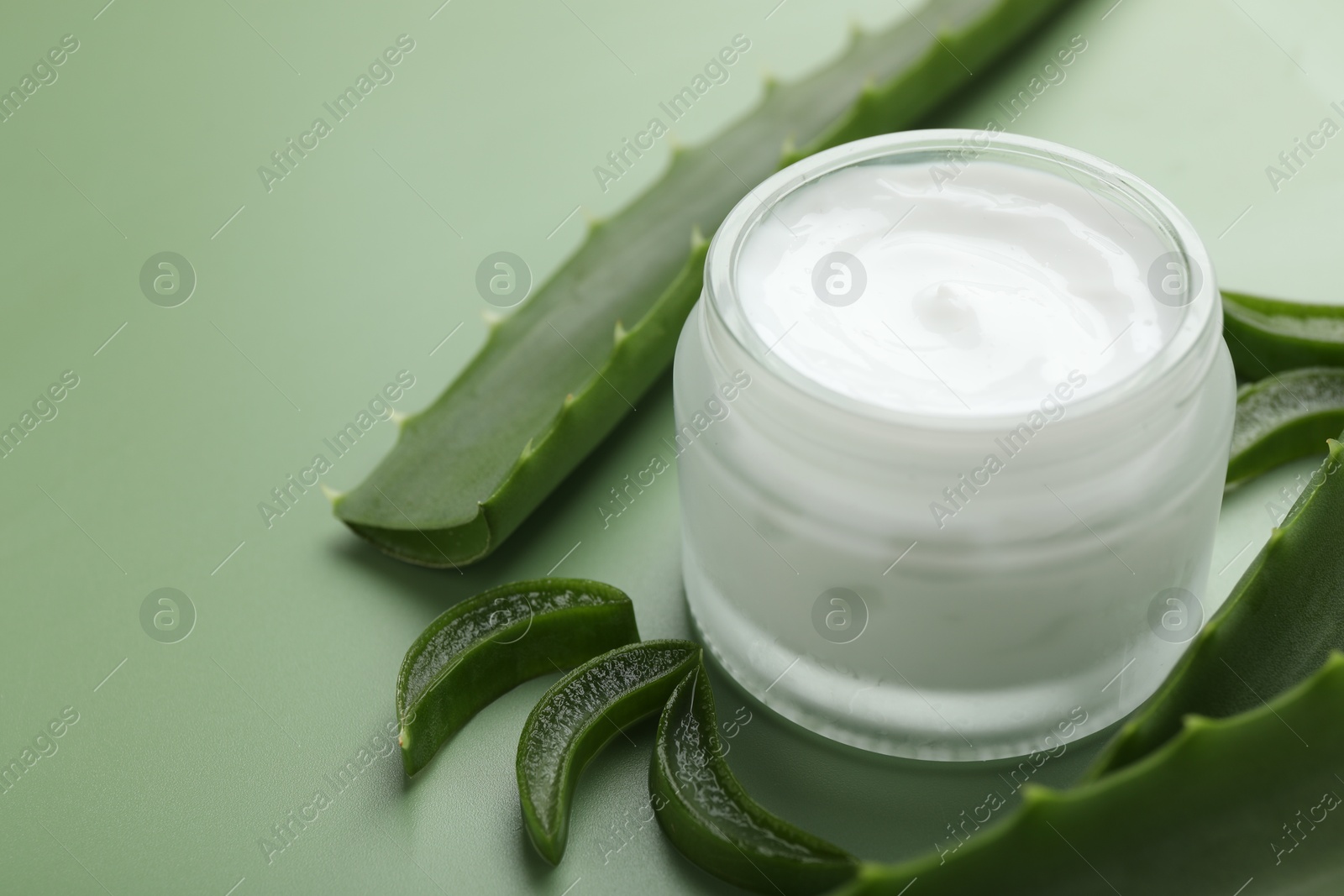 Photo of Moisturizing cream with extract of aloe vera in jar and cut leaves on green background, closeup. Space for text