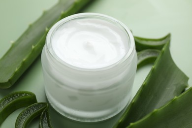 Moisturizing cream with extract of aloe vera in jar and cut leaves on green background, closeup