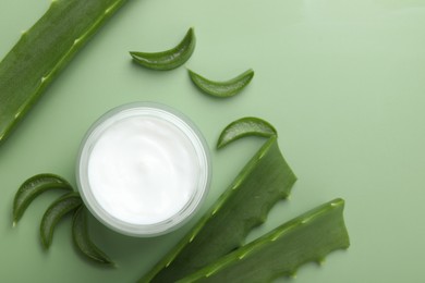 Photo of Moisturizing cream with extract of aloe vera in jar and cut leaves on green background, flat lay