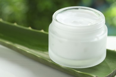 Moisturizing cream with extract of aloe vera in jar and cut leaves on white wooden table against blurred green background, closeup