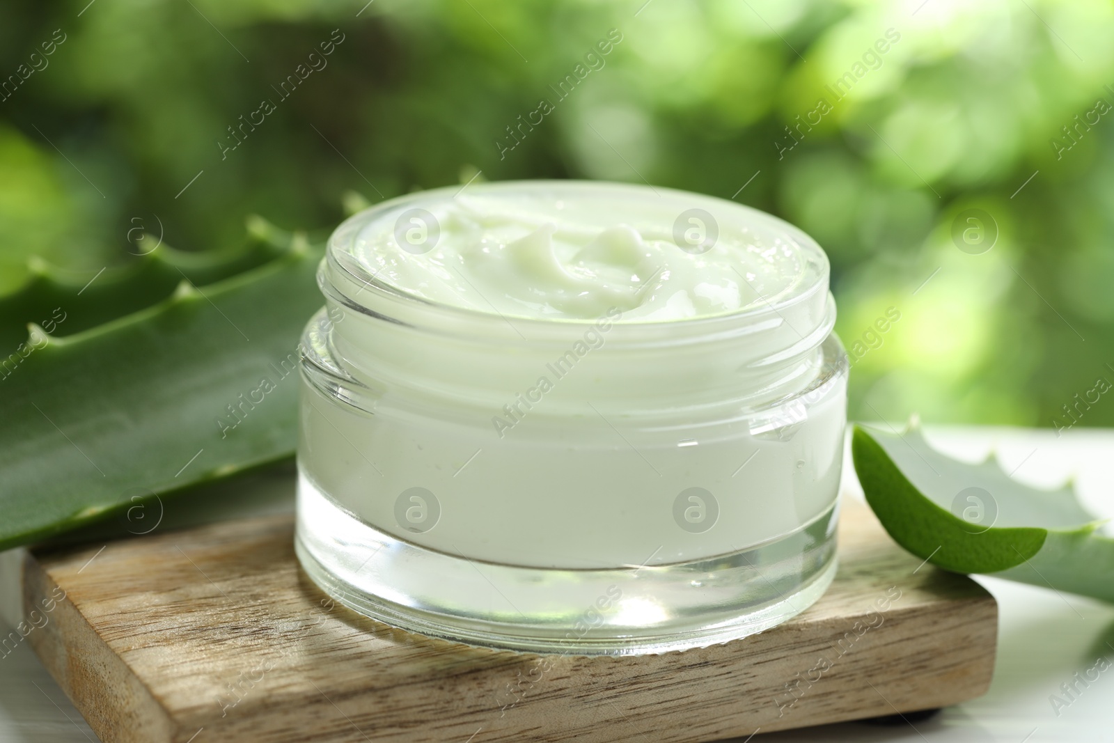 Photo of Moisturizing cream with extract of aloe vera in jar and cut leaves on white table against blurred green background, closeup