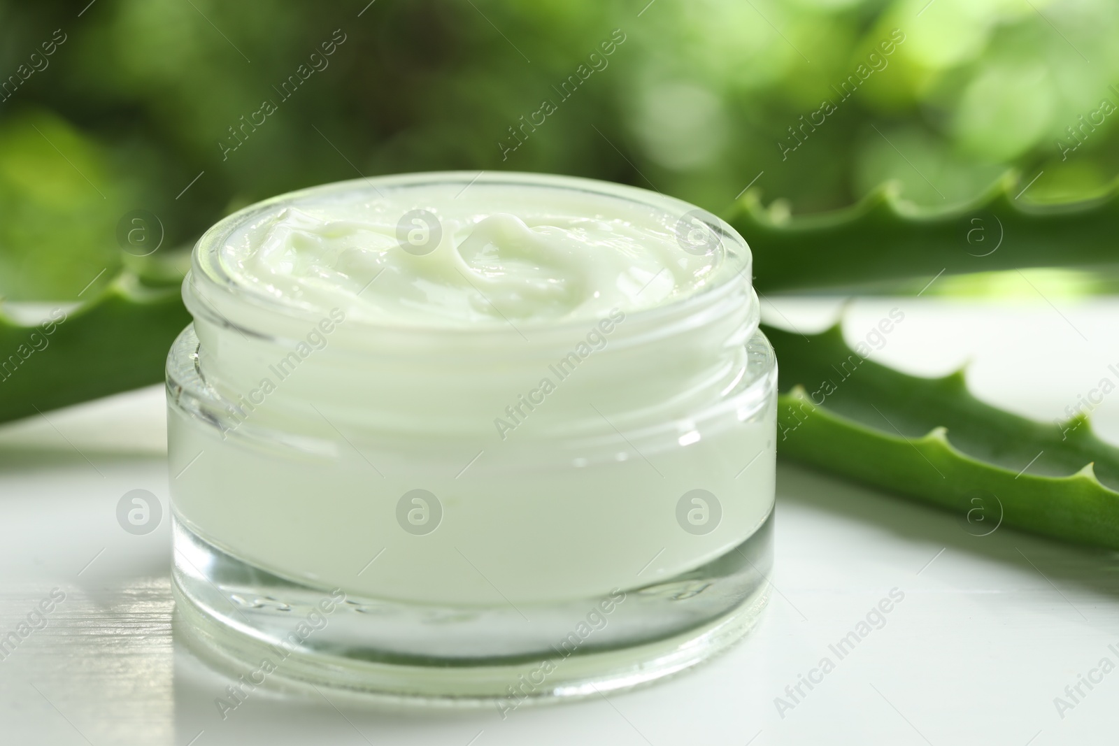 Photo of Moisturizing cream with extract of aloe vera in jar and cut leaves on white wooden table against blurred green background, closeup