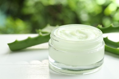 Moisturizing cream with extract of aloe vera in jar and cut leaves on white wooden table against blurred green background, closeup