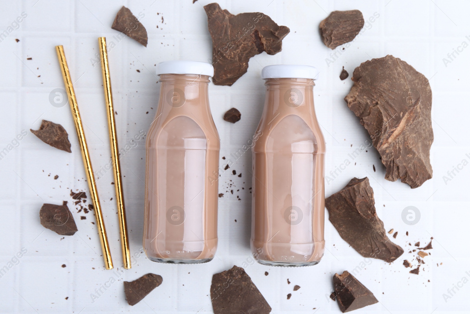 Tasty chocolate milk in glass bottles, straws and pieces of chocolate on white tiled table, flat lay Photo of Tasty chocolate milk in glass bottles, straws and pieces of chocolate on white tiled table, flat lay