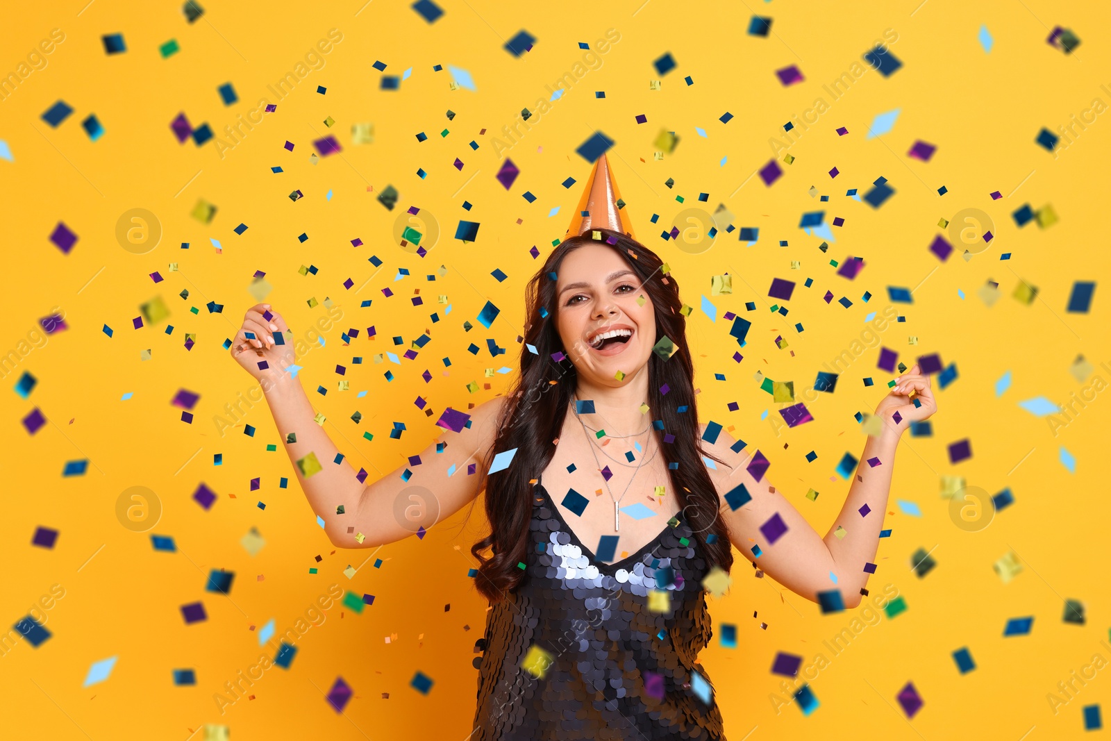 Image of Happy woman in conical paper hat under falling confetti on orange background. Surprise party